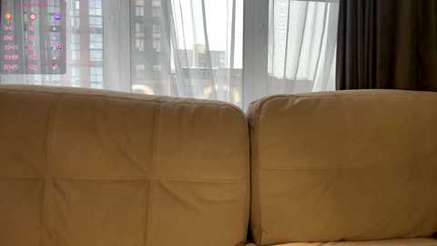 Video of a beige leather couch with a sunlit window in the background, showing an urban cityscape with high-rise buildings and a cloudy sky.