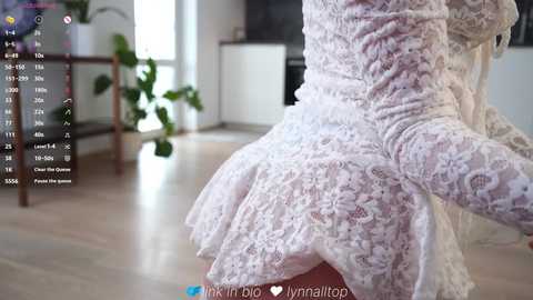 Video of a person in a white lace dress, viewed from behind, showing a curvy figure with a prominent, rounded buttocks. Background features a modern, light-filled room with plants and a wooden table.