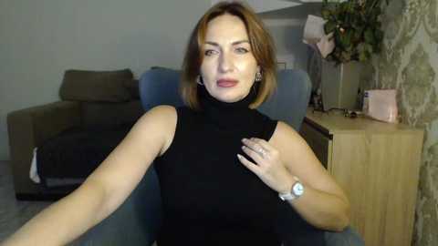 Video of a woman with light skin, shoulder-length brown hair, wearing a black sleeveless turtleneck, seated in a blue armchair, indoors.