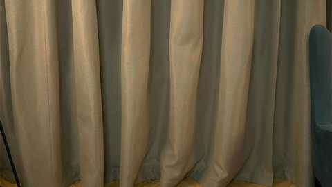 Video of beige, pleated curtains with a slightly shiny texture, drawn to one side, revealing a soft blue wall and part of a wooden floor.