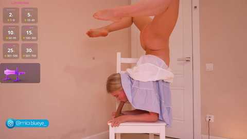 Video of a fit woman doing a handstand on a white chair, wearing a light blue shirt, in a beige-walled room with a fitness app interface displaying workout stats.