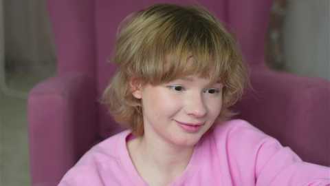 Video of a young girl with fair skin and light brown, wavy hair, wearing a pink V-neck shirt, smiling, sitting in a pink upholstered chair.