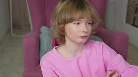 A video of a young Caucasian girl with light blonde hair, wearing a pink sweater, sitting in a pink armchair, indoors with soft, pastel-colored decor.