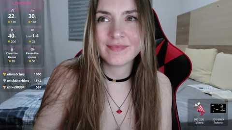Video of a smiling, fair-skinned woman with long, straight brown hair, wearing a black choker and a red necklace, sitting in a red and black gaming chair in a bedroom with a bed and window.