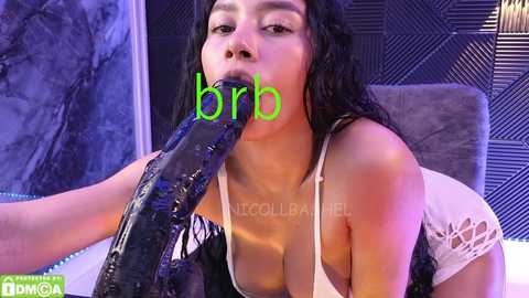 Media: Video of a woman with long black hair, wearing a white lace lingerie top, performing oral sex on a large, black, shiny dildo. Background features a dark, geometric-patterned wall and a green logo.