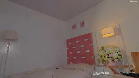 Video of a modern bedroom with a white bed, pink wall-mounted shelf, floral arrangement, beige lamp, and a white ceiling.