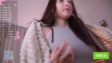 A video of a young woman with long dark hair, wearing a gray top and a white fuzzy blanket, standing indoors with a green DMCA watermark overlay.