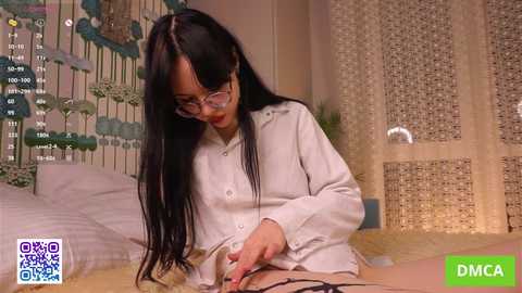 A video of a slim, fair-skinned woman with long black hair and glasses, wearing a white shirt, masturbating on a bed with a white bedspread and green floral wallpaper.