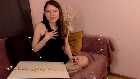 Media: Video of a smiling young woman with long brown hair, wearing a black dress, sitting on a pink couch with gold pillows and string lights, holding a large white box with gold text.