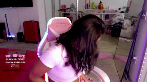 A video of a woman with long black hair in a white gaming chair, wearing a white top, in a messy room with a red suitcase, a TV, and a desk.