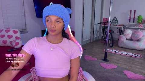 A video of a young woman with dark skin and long braids wearing a blue beanie and pink crop top, posing in a colorful bedroom with pink heart pillows and a \"Pandora\" doll.
