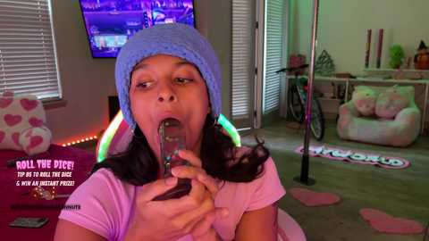 Video of a young woman with medium brown skin, wearing a light blue knit hat, purple shirt, and eating a spoonful of ice cream in a cozy, colorful bedroom.