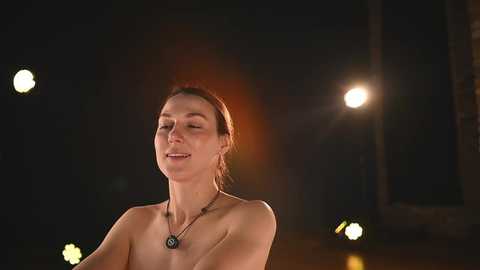 A video of a topless woman with fair skin and auburn hair, wearing a necklace, standing in dimly lit room with soft lighting.