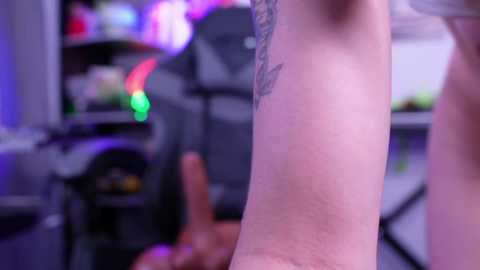 Video of a tattooed arm with a large black butterfly design, viewed from a close-up angle, set in a colorful, modern gaming room with neon lights, computer desk, and gaming chair in the background.