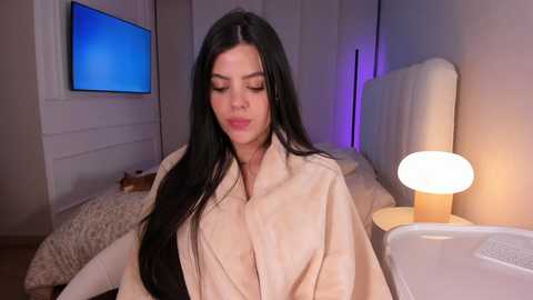 Video of a woman with long, straight black hair, wearing a beige robe, sitting on a bed in a modern, dimly lit bedroom. A round bedside lamp and a TV are visible in the background.
