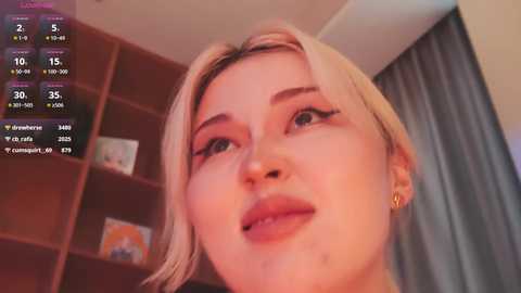 A close-up video of a young, fair-skinned woman with blonde hair, wearing a subtle makeup look, looking directly at the camera.