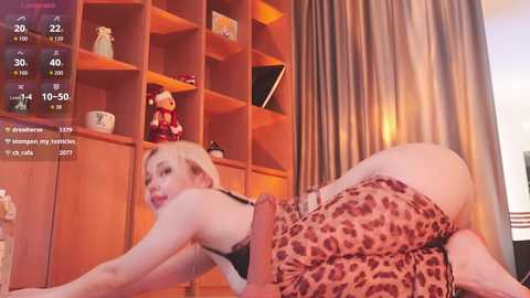Media: Video of a blonde woman in leopard print shorts, lying on her stomach on a wooden floor, with a wooden shelf in the background.
