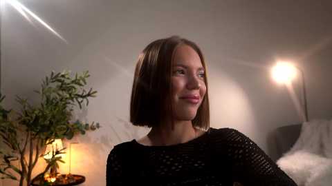 Video of a smiling Caucasian woman with short brown hair, wearing a black, textured dress, standing in a softly lit room with greenery and a glowing lamp.