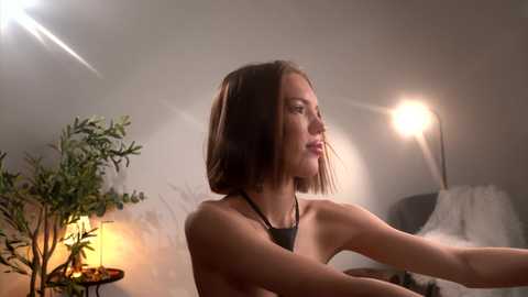 A video of a young woman with shoulder-length brown hair, wearing a black top, standing in a dimly lit room with a potted plant and a spotlight illuminating her face.