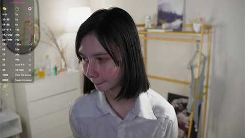 A video of a young woman with straight, shoulder-length black hair, wearing a white button-up shirt, standing in a softly lit bedroom with a white dresser, a lamp, and a coat rack in the background.