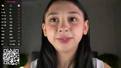 Video of a young Latina woman with straight black hair, medium skin tone, and light makeup, wearing a sleeveless top. Background shows a blurred indoor setting with a green plant. QR code in bottom left corner.