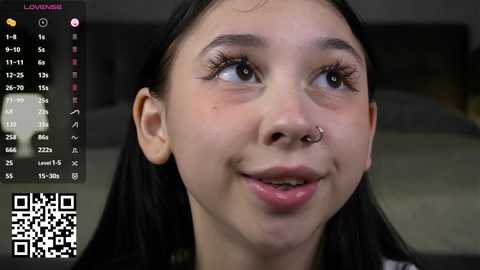 A close-up video of a young woman with fair skin, dark hair, and a silver nose ring, looking up thoughtfully. Background shows a blurred room with a digital clock and QR code.