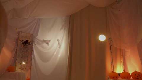 Video of a Halloween-themed room with white draped fabric, a glowing pumpkin lantern, and a spider decoration, creating a spooky ambiance.