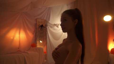 Video of a topless, slender Asian woman with long hair, covering her breasts, in a dimly lit, draped room with warm, orange lighting, candles, and a mirror.