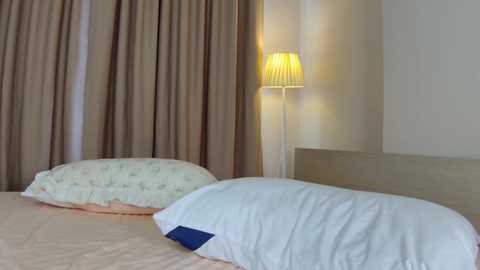 A video of a cozy bedroom with beige curtains, a light-colored headboard, a white pillow, and a lit lamp casting a warm glow.