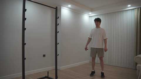 Video of a young man with short dark hair in a white T-shirt, grey shorts, and black shoes standing in a dimly lit, plain white room with a vertical pull-up bar and wall-mounted lights.