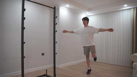 Video of a Caucasian man with short brown hair, wearing a white T-shirt, beige shorts, and black socks, performing a static stretch on a black pull-up bar in a minimalist, well-lit room with white walls and wooden floors.