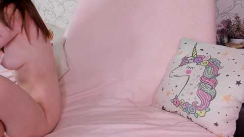 Video of a naked woman with light skin and brown hair, sitting on a bed with a pastel pink duvet. She holds a pink pillow with a unicorn design.