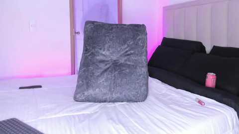 Video of a modern bedroom with a gray, plush pillow on a white bed, black headboard, and a pink can beside a remote control on a white nightstand.