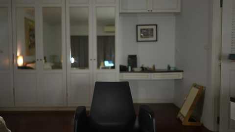 Media: Video of a dimly lit, minimalist bedroom with a black chair facing a white closet and desk area. The room features a framed artwork on the wall and scattered artwork supplies on the floor.
