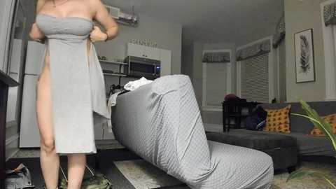 Video of a light-skinned woman in a strapless, gray dress, lifting it to reveal her legs, standing in a modern living room with a gray couch, cushions, and a recliner.