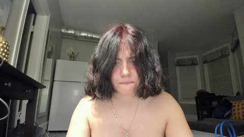 Video of a fair-skinned woman with shoulder-length, dark curly hair, topless, sitting indoors. The background features a kitchen with white appliances, blinds, and a cluttered living area.