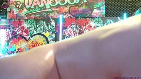 Media: Video of a hand with a tattoo, foreground, against a vibrant, colorful graffiti-covered wall in the background. The wall features abstract shapes and text, with a prominent \"VANCOUVER\" sign.