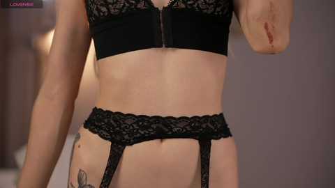 Media: Video of a woman wearing black lace lingerie, including a bra, garter belt, and panties, showcasing a slim physique with tattoos on her hips. The background is blurred, featuring neutral tones.