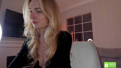 Media: A video of a fair-skinned, blonde-haired woman with wavy hair, wearing a low-cut black top, sitting indoors with a TV and window in the background.