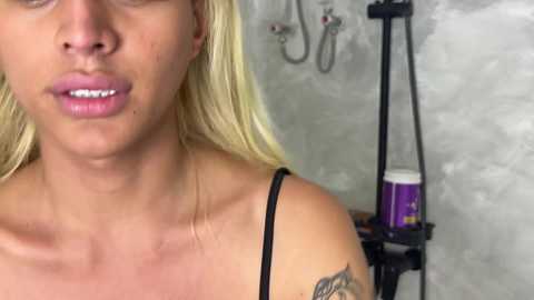 Video of a blonde woman with fair skin, wearing a black tank top, showing a tattoo on her right shoulder. She has a neutral expression, and the background features a shower with a purple shampoo bottle.