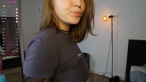 Video of a fair-skinned woman with shoulder-length brown hair, wearing a black shirt, standing in a dimly lit room with a TV and a lamp.