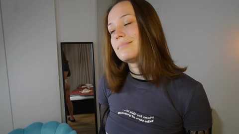 Video of a Caucasian woman with straight, shoulder-length brown hair, wearing a dark gray long-sleeve shirt with white text. She's smiling, standing indoors next to a mirror that reflects a bed with red sheets and a person in a white dress.