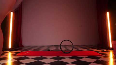Video of a dimly lit, minimalist room with black-and-white checkered floor, red yoga mat, and two vertical red curtains. A black yoga wheel rests on the mat.