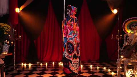 Media: Video of a vibrant, colorful Day of the Dead altar featuring a large, intricately decorated sugar skull, surrounded by flickering candles and festive decorations, set against a dark, red-draped backdrop.