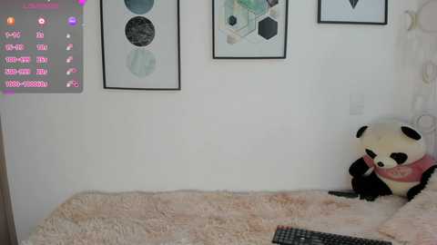 A video of a cozy bedroom with white walls, framed abstract art, a plush pink blanket, a stuffed panda bear, and a computer monitor displaying a purple interface.