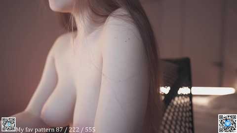 Media: Video of a topless woman with pale skin, long brown hair, and medium-sized breasts, captured from the side, in a dimly lit room with a black chair and a lamp.