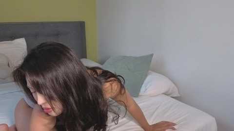 Video of a young woman with long, dark hair, slender physique, and light skin, kneeling on a bed with white sheets, pale blue pillow, and gray headboard, in a minimalistic, softly lit room.