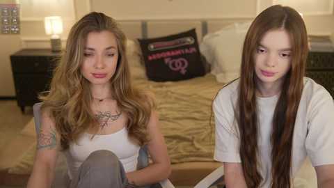 Video of two young women with long hair, one with tattoos, sitting on a bed in a cozy bedroom with beige walls and a dark wooden nightstand.