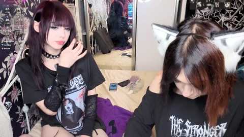 Video of two young women with dark hair, one in a black metal t-shirt, the other in a black off-shoulder shirt with cat ears, sitting in a messy bedroom with purple bedding and a mirror.