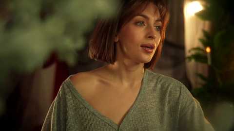 Video of a middle-aged Caucasian woman with short auburn hair, wearing a green off-shoulder sweater, outdoors in a dimly lit, softly blurred garden setting.